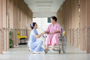 This image depicts a nurse kneeling and holding the hands of a smiling patient seated in a wheelchair. The scene takes place in a brightly lit corridor with railings on both sides. The nurse is dressed in light blue scrubs, while the patient is wearing pink attire, indicating a caring and supportive interaction in a healthcare setting