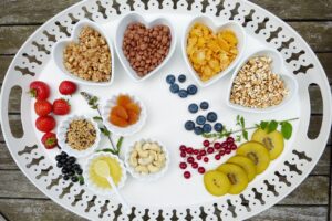 A decorative white tray filled with a variety of healthy foods, including heart-shaped bowls of granola, chocolate cereal balls, cornflakes, and puffed grains. Surrounding these bowls are small dishes of dried apricots, cashews, honey, and mixed seeds. Fresh fruits such as strawberries, blueberries, red currants, black currants, and sliced kiwi are also arranged on the tray, creating a vibrant, nutritious assortment.